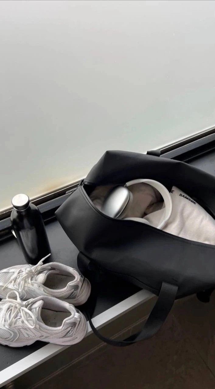 A black leather duffel bag sits open on a windowsill next to a black bottle and a pair of white sneakers, with a white shoebox partially visible inside the bag.