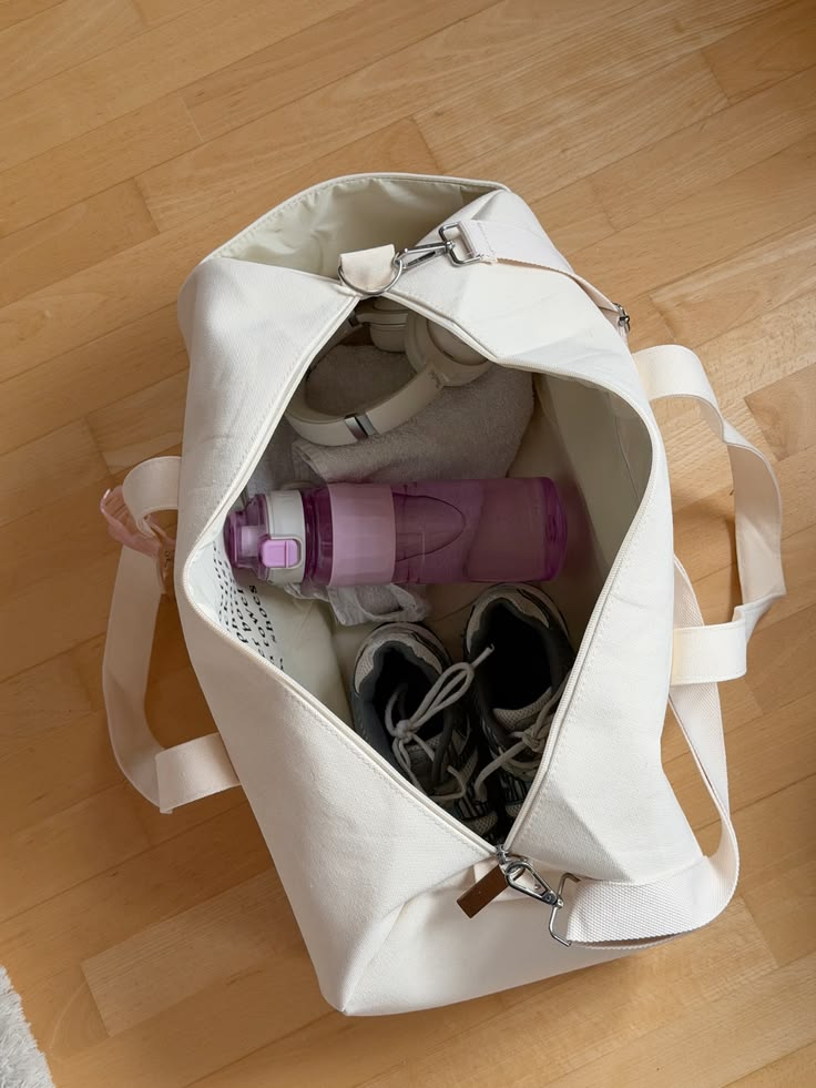 An off-white canvas duffel bag filled with gym items, including a purple water bottle, a gray sweatshirt, and a pair of black and white sneakers, placed on a light wood floor.